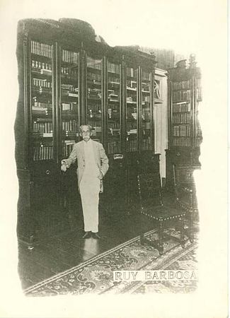 Rui Barbosa em 1910 (?) em pé junto às estantes da biblioteca em sua residência, hoje "Sala Constituição" do atual Museu Casa de Rui Barbosa. Original p/b, com legenda: "RUY BARBOSA".